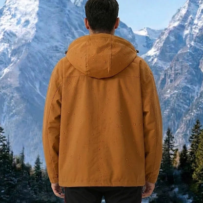 Vue dos veste coloris caramel — capuche, gouttes d'eau visibles sur le tissu, fond montagne et forêt de pins